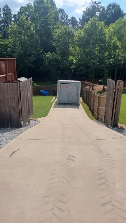 A STORsquare portable storage container positioned at the bottom of a long, steep concrete driveway between wooden fences and trees.