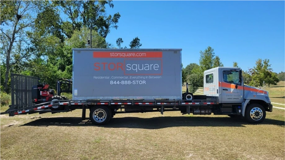 STORsquare moving container secured on a flatbed truck for transport across a grassy Memorial property.